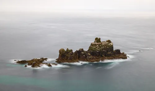 This landscape photograph captures a long exposure view of rugged rocks rising from the sea off the coast of Cornwall. The image shows the tranquil surface of the water, smoothed out by the long exposure technique, with the jagged cliffs and rocky outcrops contrasted against the gentle hues of the evening sky. Taken in early summer, the light suggests late afternoon approaching evening, illuminating the textures of the cliffs and the surrounding turquoise sea. The scene emphasizes the geological character of the Cornish coastline, with the isolated rocky formations standing prominently as the main subject in the composition.